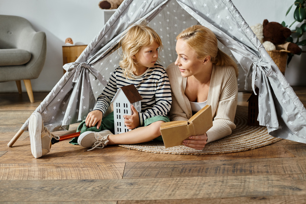 маленькая девочка с протезной ногой и блондинка мать читает книгу и сидит в игровой палатке дома - Фото, изображение
