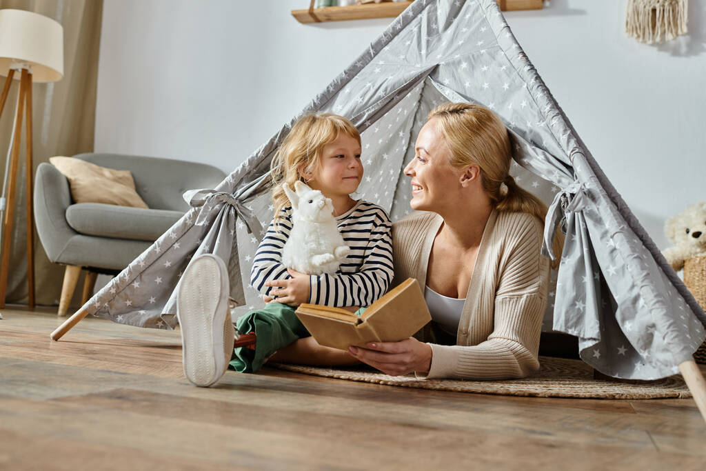 улыбающаяся девушка с протезной ногой, держащая мягкую игрушку рядом с матерью, читающей книгу в детской палатке дома - Фото, изображение