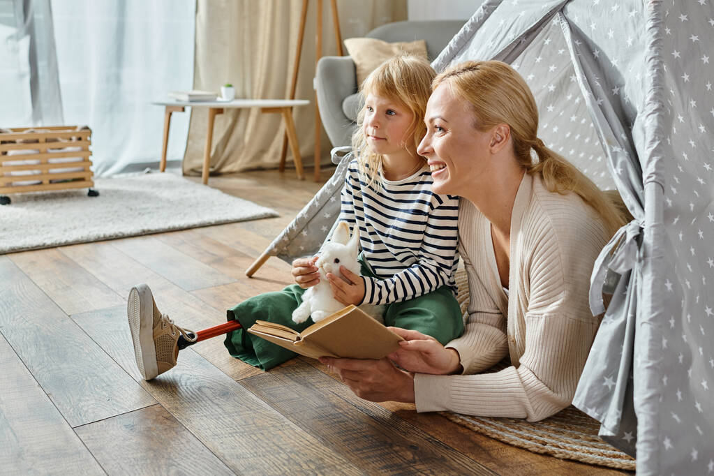мечтательная девушка с протезной ногой, а мать смотрит в сторону и читает книгу, сидя в игровой палатке - Фото, изображение
