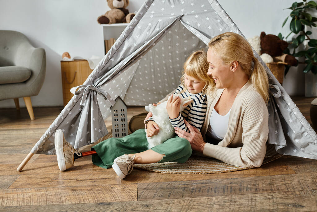 niña con prótesis pierna abrazando suave juguete conejo cerca feliz madre mientras está sentado en juego tienda de campaña - Foto, Imagen