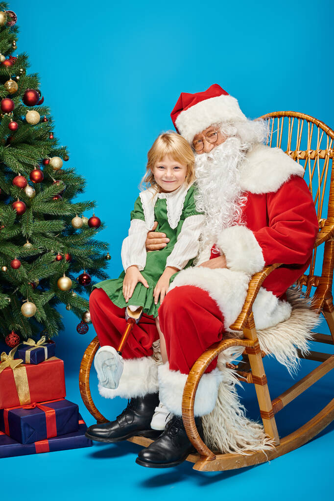 happy girl with prosthetic leg sitting on laps of Santa Claus next to Christmas tree on blue - Photo, Image