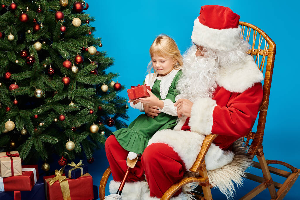 щаслива дівчина з протезною ногою сидить на кільцях Діда Мороза з подарунком поруч з ялинкою - Фото, зображення