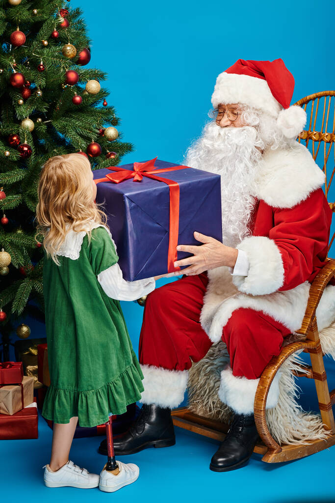 Santa Claus giving present to happy girl with prosthetic leg next to Christmas tree on blue backdrop - Photo, Image