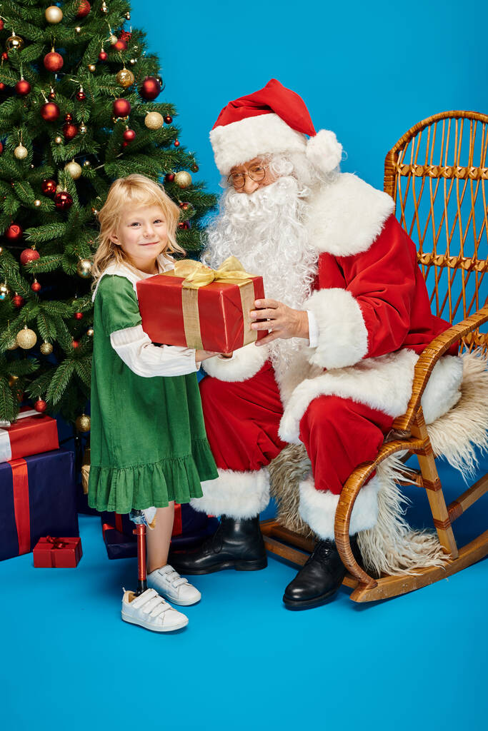 Père Noël donnant cadeau à une fille heureuse avec la jambe prothétique à côté de l'arbre de Noël sur fond bleu - Photo, image