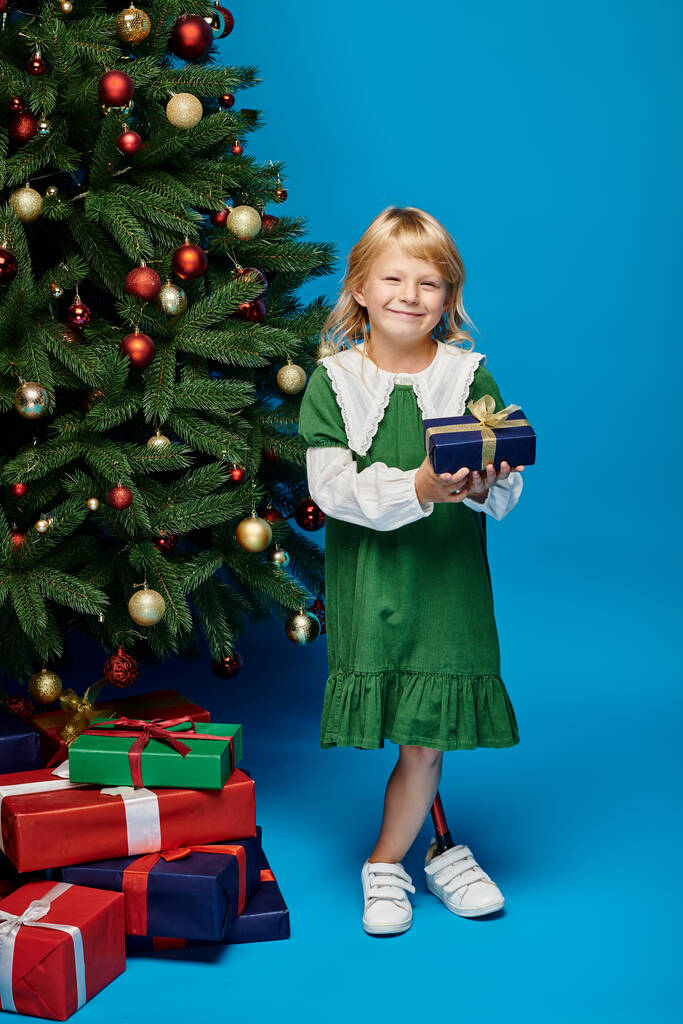 gelukkig meisje in jurk met kunstbeen houden verpakt cadeau naast kerstboom op blauw - Foto, afbeelding