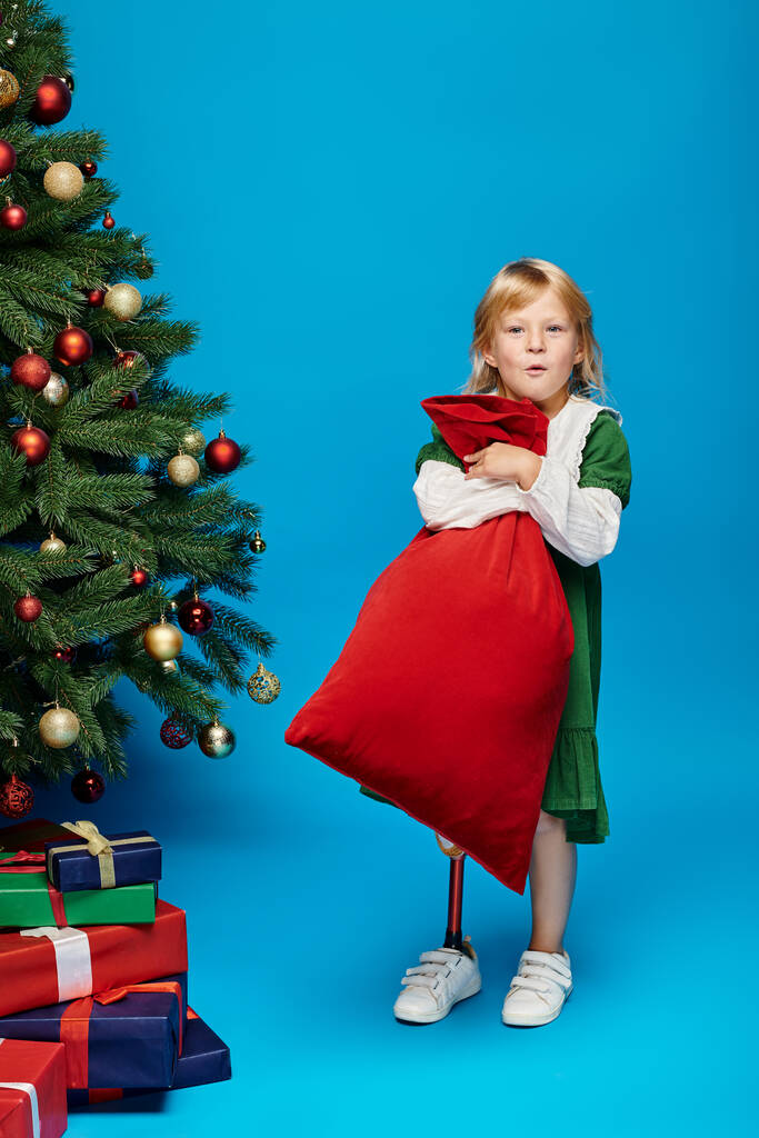 amazed girl in dress with prosthetic leg holding sack bag with presents near Christmas tree on blue - Photo, Image