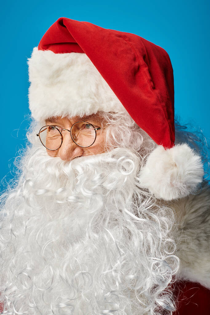 portrait of happy Santa Claus with white beard and eyeglasses looking away on blue background - Photo, Image