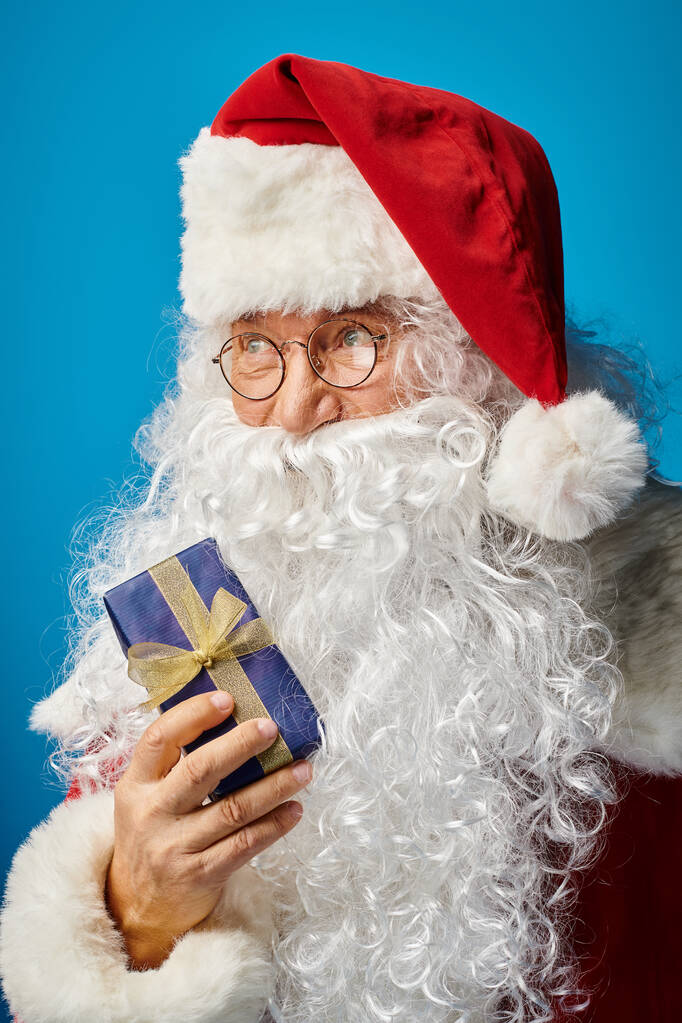 портрет счастливого Санта-Клауса с белой бородой и очками, держащих рождественский подарок на голубом - Фото, изображение