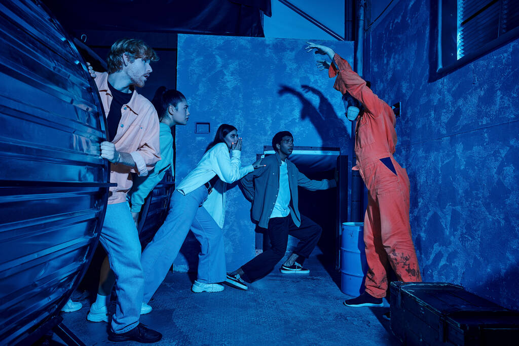 Πολυπολιτισμική ομάδα ανθρώπων που δραπετεύουν από τον άνθρωπο με μάσκα αερίου κατά τη διάρκεια περιπέτειας στην αίθουσα αναζήτησης - Φωτογραφία, εικόνα