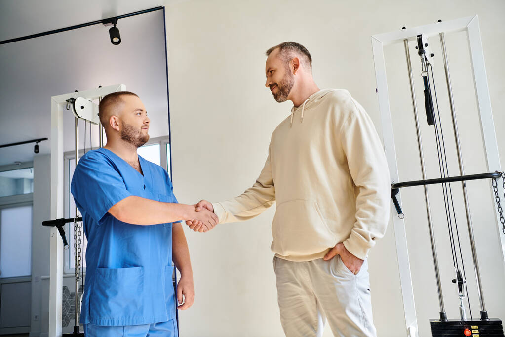 Улыбающийся мужчина пожимает руку молодому доктору в синей форме в реабилитационном центре Кинезио - Фото, изображение