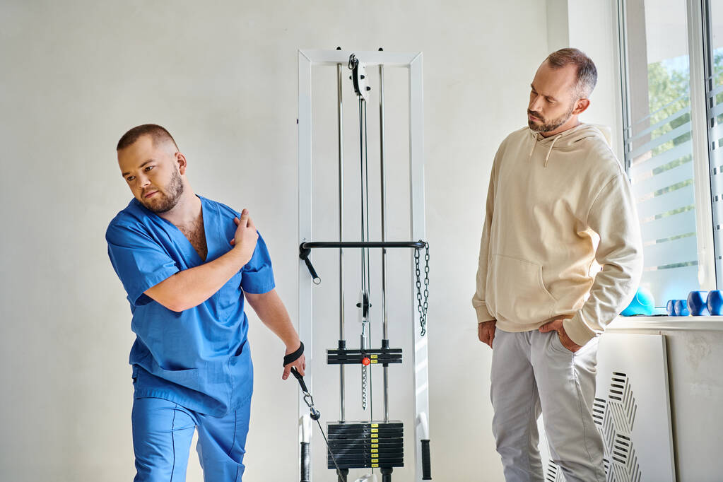 фізіотерапевт у блакитній формі навчає чоловіка біля тренувальної машини в реабілітаційному центрі - Фото, зображення