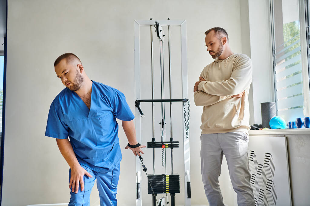 mladý lékař v modré uniformě poučení muže v blízkosti tréninkového stroje v rehabilitačním centru - Fotografie, Obrázek