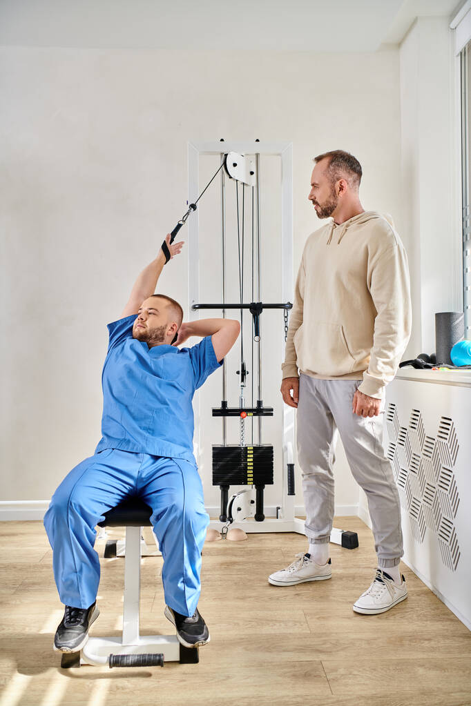 treinamento de fisioterapeuta jovem na máquina de exercício durante consulta do homem no centro de kinesio - Foto, Imagem
