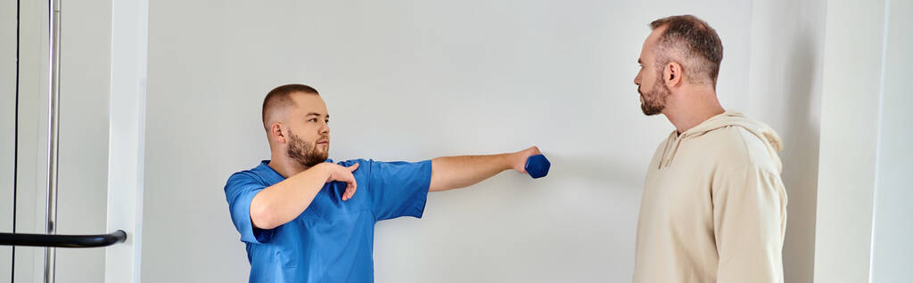 Mavi üniformalı doktor, Kinesio merkezinde dambılla egzersiz yapıyor, yatay pankart. - Fotoğraf, Görsel