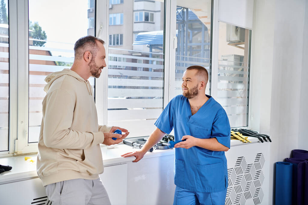 jeune spécialiste en uniforme bleu parlant à son patient masculin dans le centre de kinesio, réadaptation - Photo, image