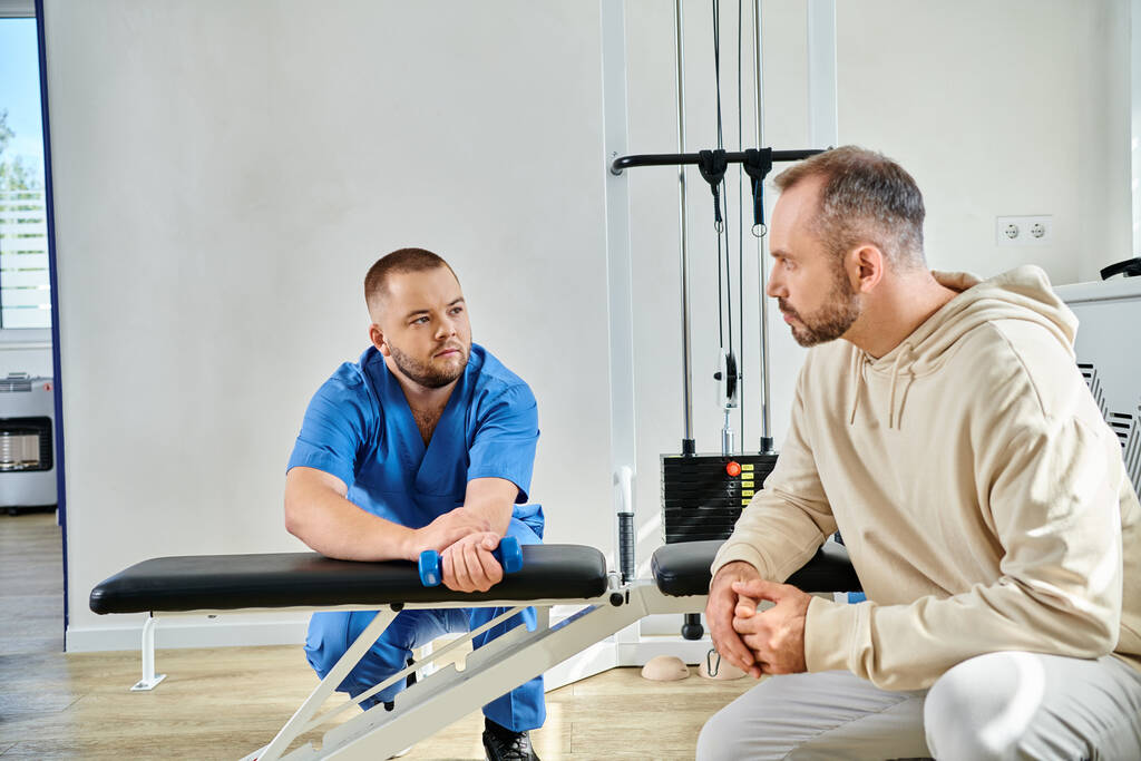mannelijke patiënt op zoek naar rehabilitoloog tonen oefening met halter in de sportschool van kinesio centrum - Foto, afbeelding