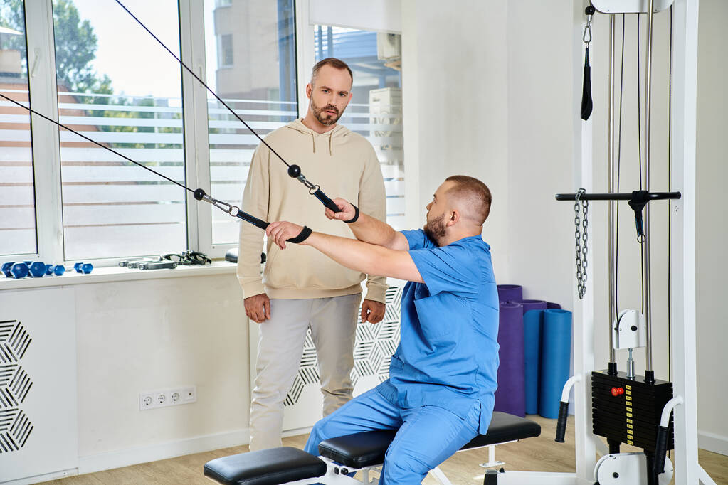 rehabilitologist in blue uniform showing exercise on training machine to man in kinesio center - Photo, Image