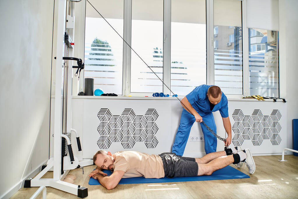 врач в синей форме помогает мужчине тренироваться на тренажере в тренажерном зале кинезио-центра - Фото, изображение