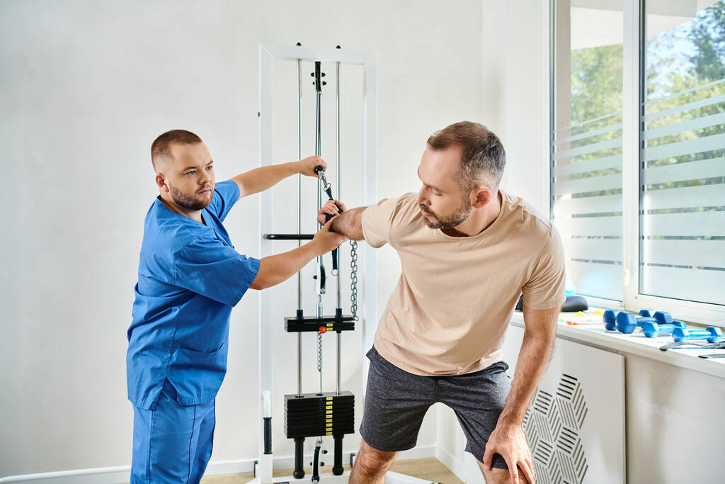 man in sportswear working out on exercise machine with help of recovery specialist in kinesio center - Photo, Image