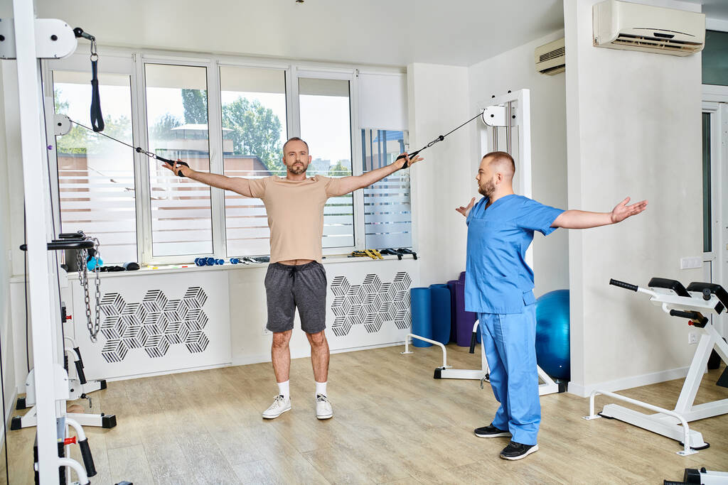 jonge fysiotherapeut consulting mannelijke patiënt uit te werken op de oefening machine in kinesio centrum - Foto, afbeelding