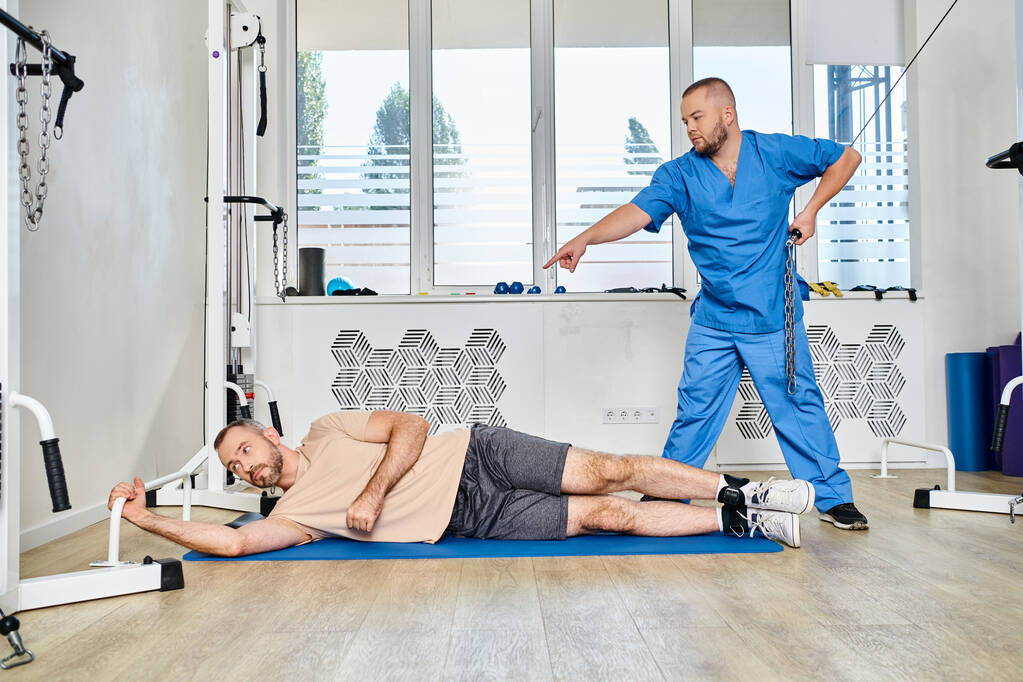 doctor in blue uniform pointing and instructing man during recovery training in kinesio center - Photo, Image