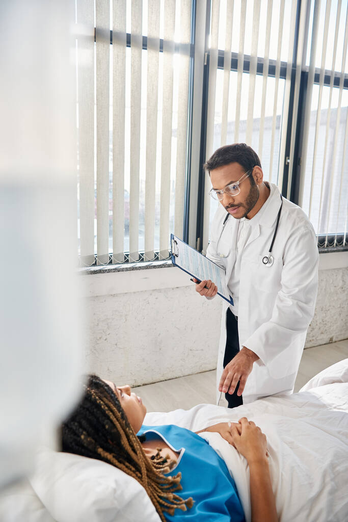 inteligentny indyjski lekarz rozmawia ze swoją afrykańską amerykańską pacjentką leżącą w łóżku na oddziale opieki zdrowotnej - Zdjęcie, obraz