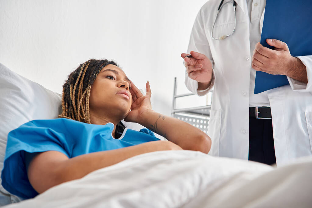 άρρωστη Αφροαμερικανή γυναίκα ξαπλωμένη στο κρεβάτι στο νοσοκομείο ακούγοντας τον νεαρό Ινδό γιατρό της. - Φωτογραφία, εικόνα