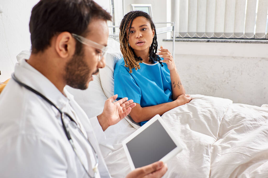 enfoque en la mujer afroamericana en la cama de hospital mirando al doctor indio borroso con la tableta - Foto, imagen