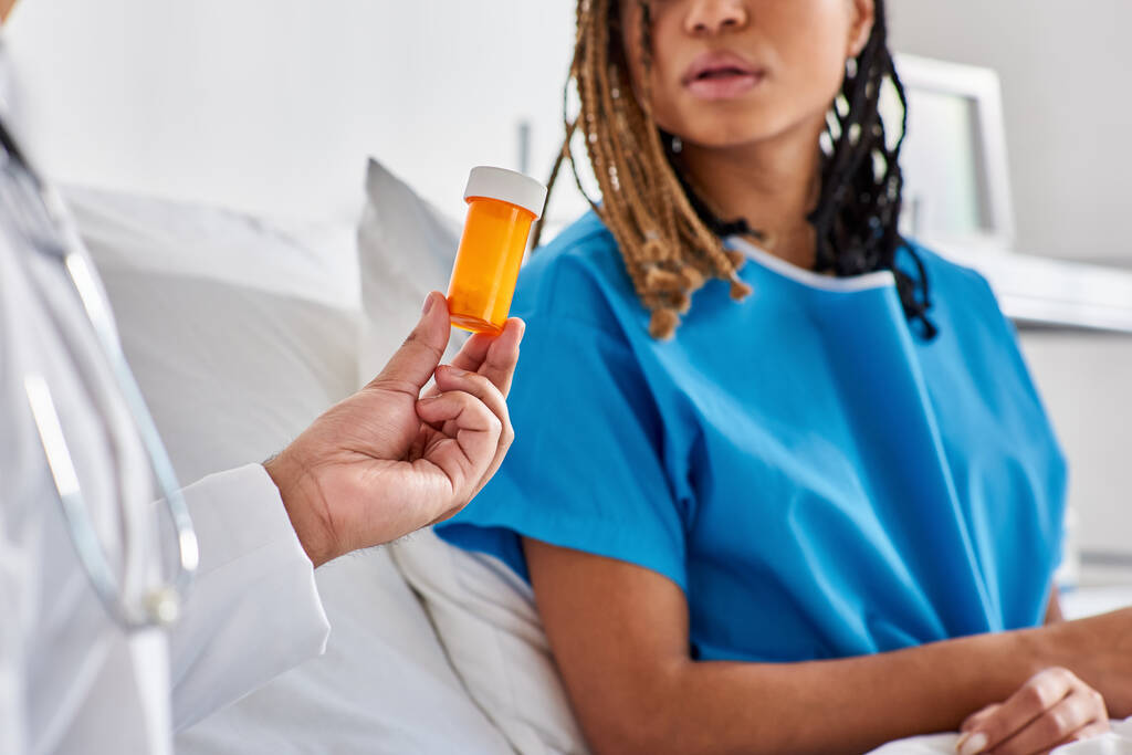 kranke afrikanisch-amerikanische Frau schaut ihren indischen Arzt an, der ihr auf ihrer Krankenhausstation Tabletten verabreicht - Foto, Bild