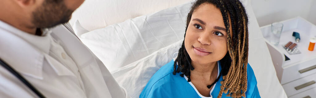 focus on jolly african american woman in bed looking calmly at her blurred indian doctor, banner - Photo, Image