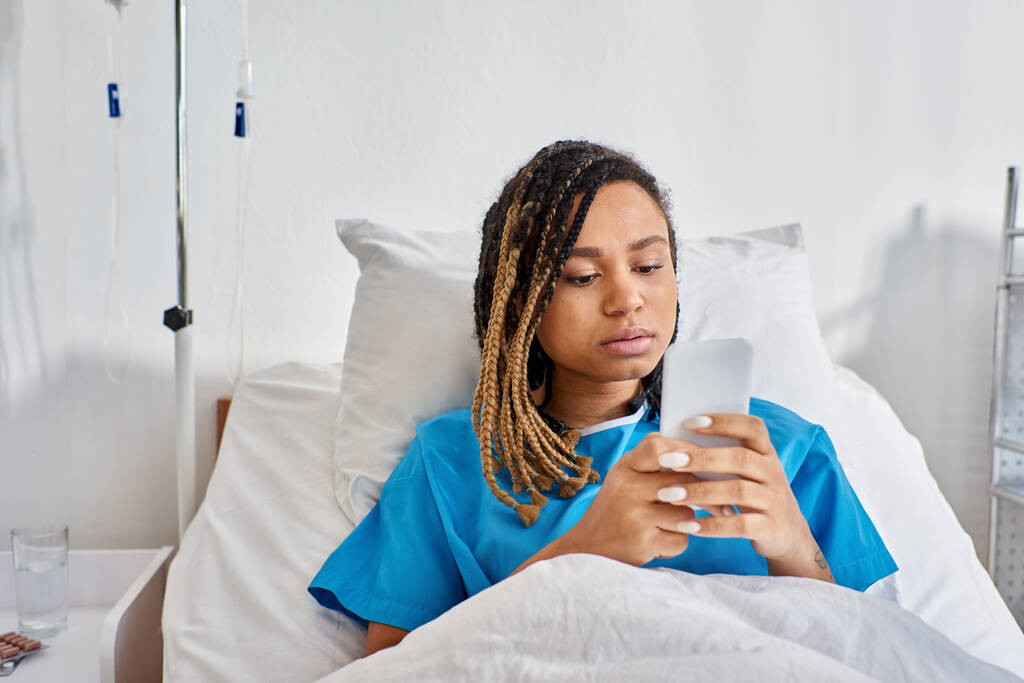 young african american woman lying in hospital bed and looking at her mobile phone, healthcare - Photo, Image