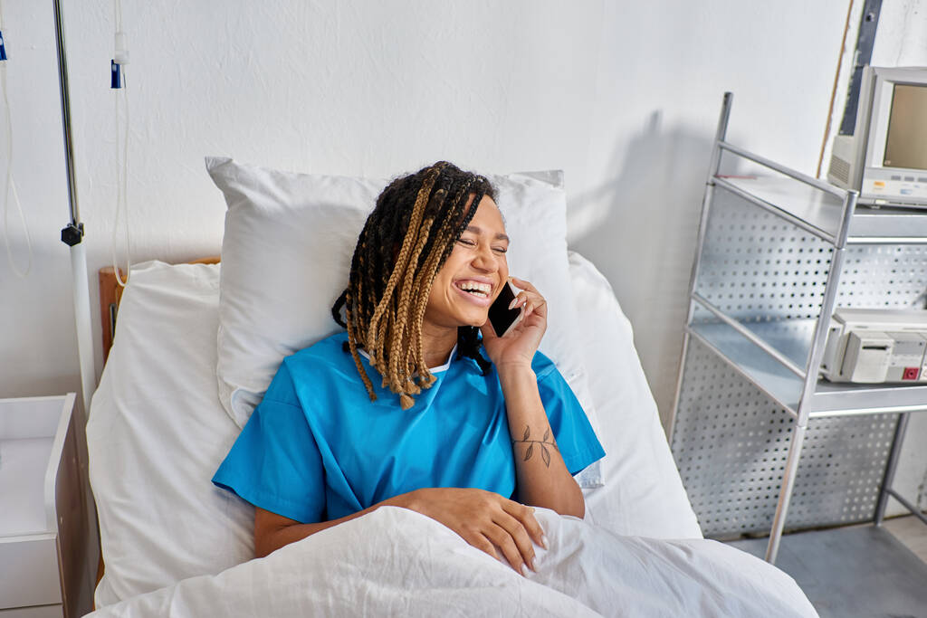 Neşeli Afrikalı genç Amerikalı kadın telefonla konuşuyor ve hastane koğuşunda gülümsüyor. - Fotoğraf, Görsel