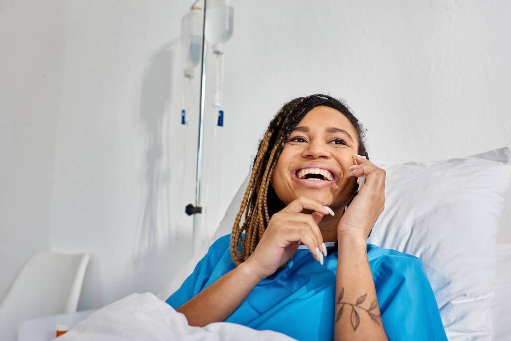 alegre joven afroamericana mujer hablando por teléfono y sonriendo en su sala de hospital, atención médica - Foto, Imagen