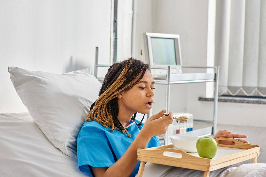 jeune femme afro-américaine ayant du porridge et de la pomme pour le petit déjeuner en salle d'hôpital, soins de santé - Photo, image
