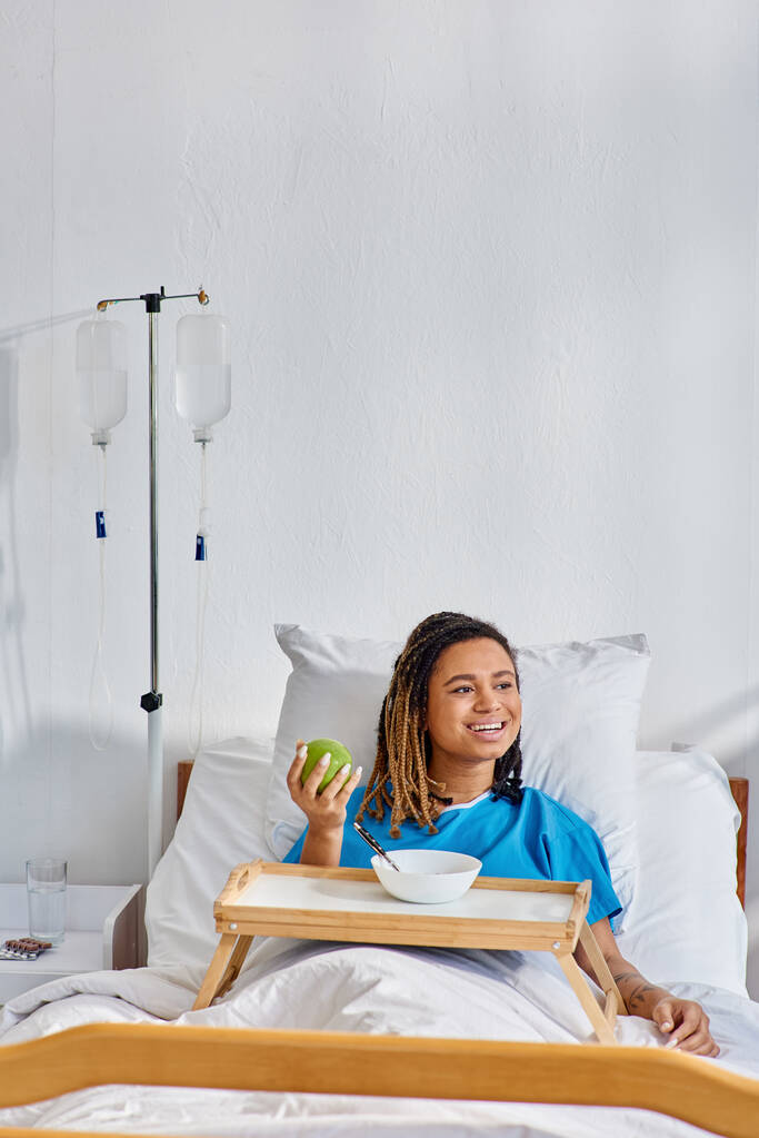 jong Afrikaans amerikaans vrouw met pap en appel voor ontbijt in ziekenhuis afdeling, gezondheidszorg - Foto, afbeelding