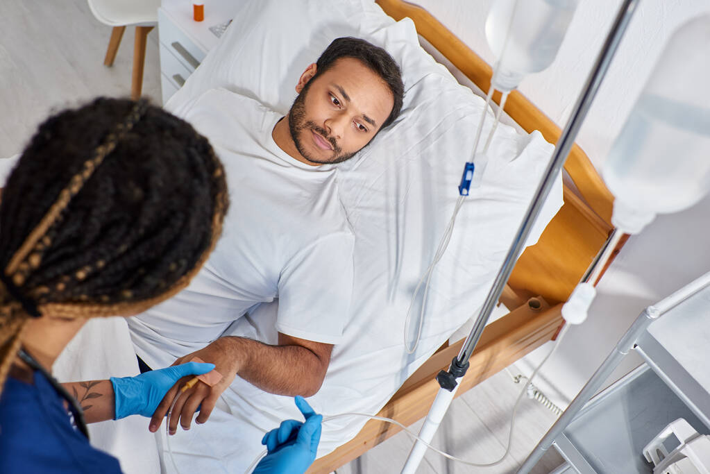foco no homem indiano deitado na cama do hospital enquanto enfermeira afro-americana definindo seu contador de gotas - Foto, Imagem