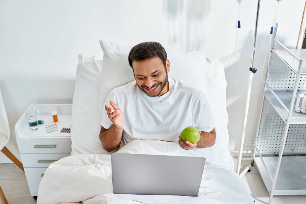 młody Indianin mając wideo rozmowy i trzymając zielone jabłko leżąc w szpitalu łóżko, opieka zdrowotna - Zdjęcie, obraz