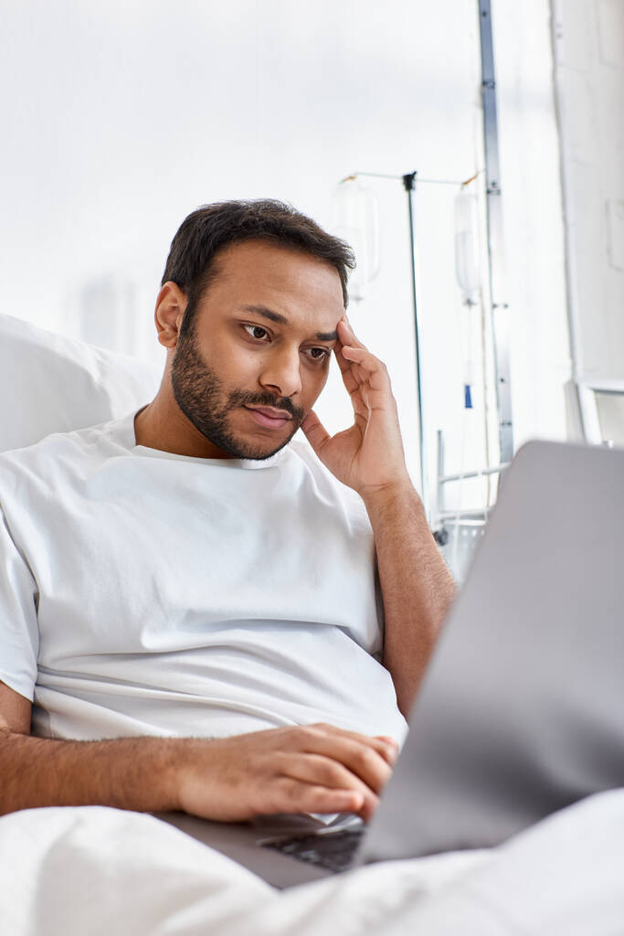 paciente indio concentrado acostado en la cama de hospital en su sala mirando a su computadora portátil, atención médica - Foto, imagen