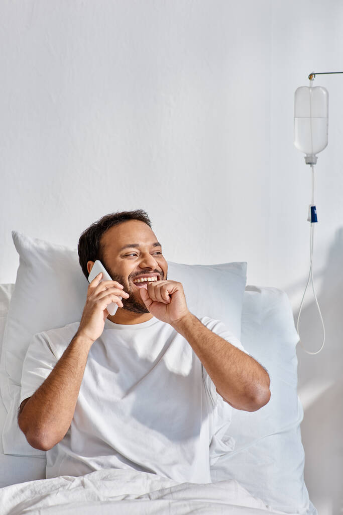 Χαρούμενος νεαρός Ινδός άνδρας που μιλάει χαρούμενα από το τηλέφωνο ενώ βρίσκεται στο κρεβάτι του σε νοσοκομειακό θάλαμο, υγειονομική περίθαλψη - Φωτογραφία, εικόνα