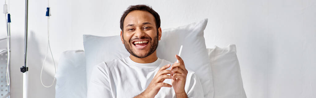 vidám indiai férfi fekszik a kórházi ágyon telefonnal a kezében és vidáman mosolyog, banner - Fotó, kép