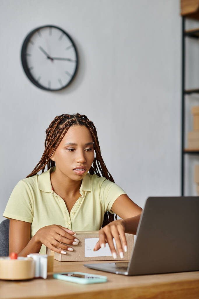gut aussehende afrikanisch-amerikanische Einzelhändlerin schaut auf ihren Laptop, während sie hart arbeitet, Lieferung - Foto, Bild