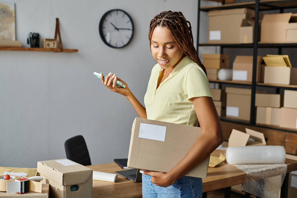 atraktivní africká americká prodavačka drží telefon a dívá se na kartonovou krabici, dodání - Fotografie, Obrázek