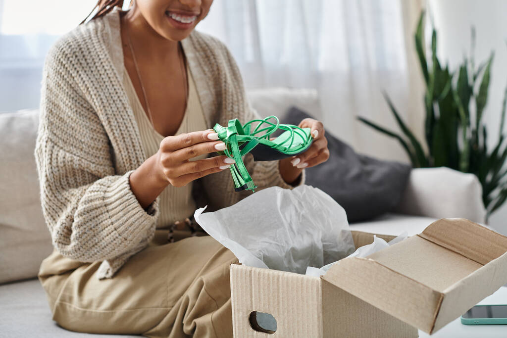 abgeschnittene Ansicht einer freudigen afrikanisch-amerikanischen Frau mit Zahnspange beim Auspacken ihrer neuen grünen Schuhe zu Hause - Foto, Bild