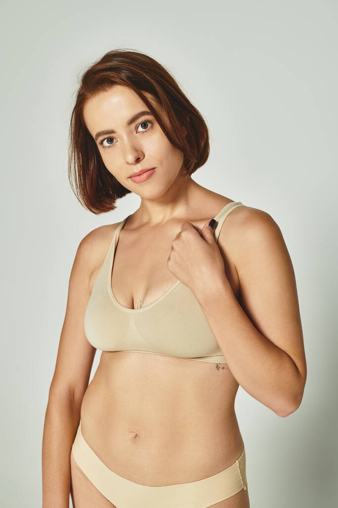 auto-confiante e jovem mulher em lingerie bege confortável olhando para a câmera no fundo cinza - Foto, Imagem