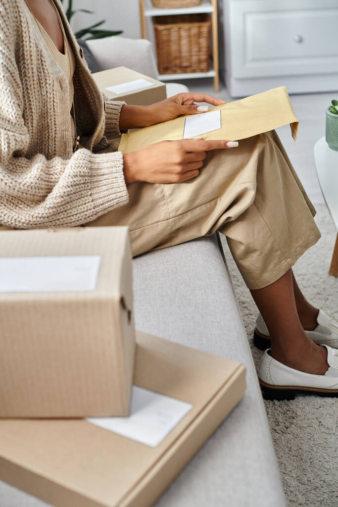 abgeschnittene Ansicht einer jungen afrikanisch-amerikanischen Frau in Homewear beim Öffnen eines Postpakets, während sie auf der Couch sitzt - Foto, Bild