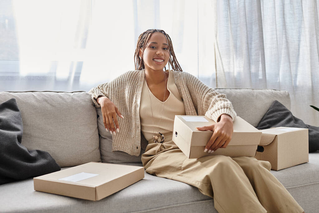 radosna młoda Afroamerykanka w przytulnym stroju, siedząca na kanapie z pudełkami i uśmiechnięta do kamery - Zdjęcie, obraz