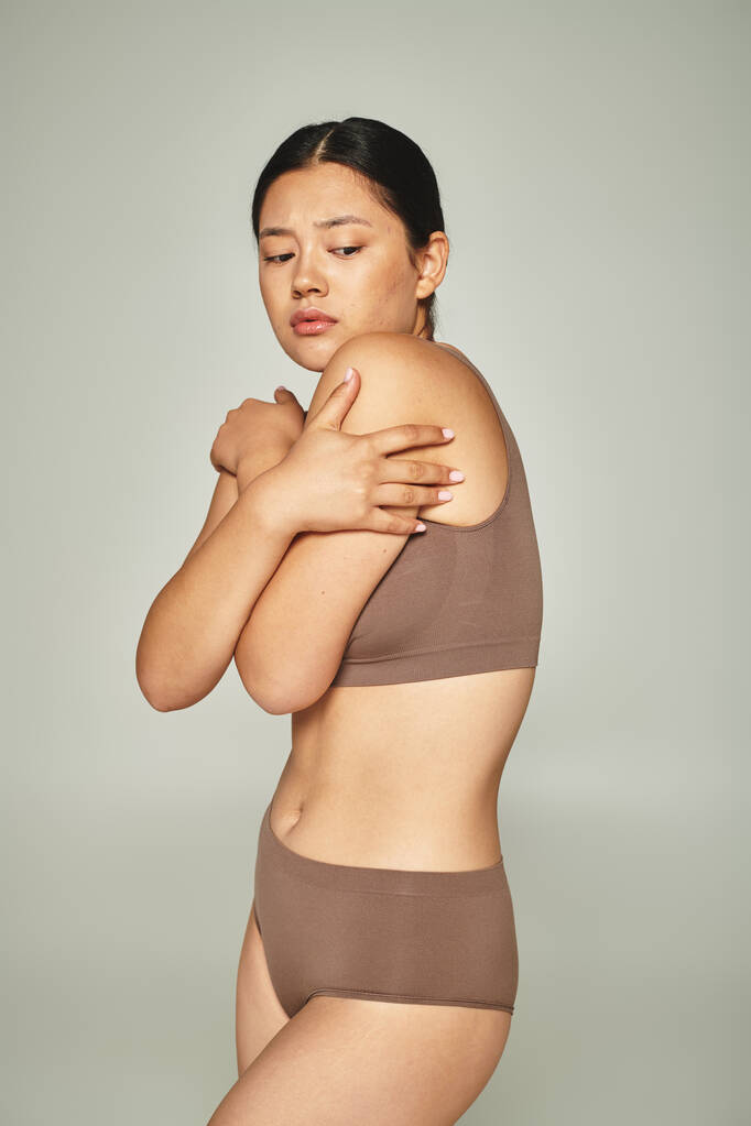 schüchterne asiatische Frau in Unterwäsche, die ihren Körper bedeckt, während sie sich auf grauem Hintergrund umarmt, body shaming - Foto, Bild