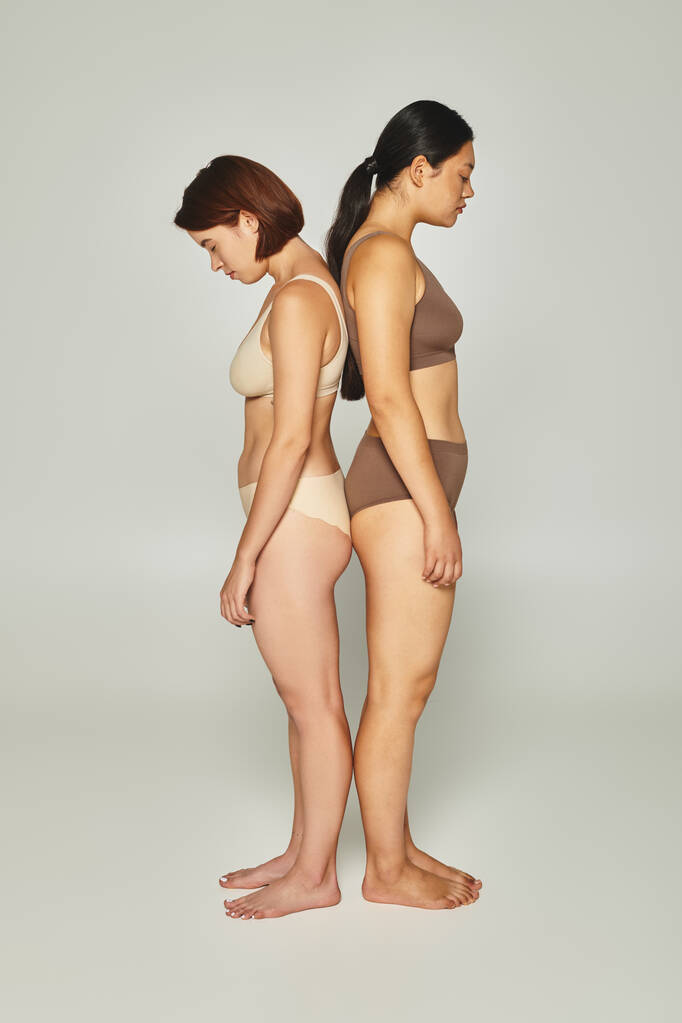 засмучені мультикультурні жінки в нижній білизні, стоячи назад на сірому тлі, сором'язливі тіла - Фото, зображення