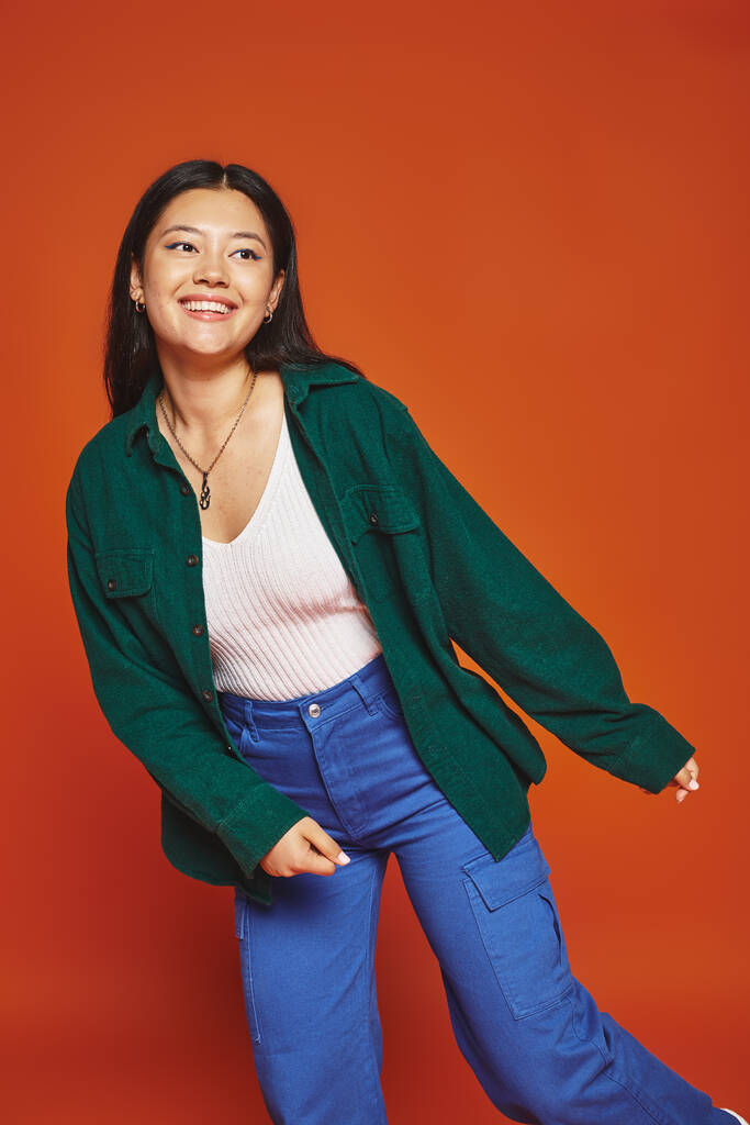fröhliche junge asiatische Frau posiert in lebendigem Outfit und schaut auf orangefarbenem Hintergrund weg - Foto, Bild