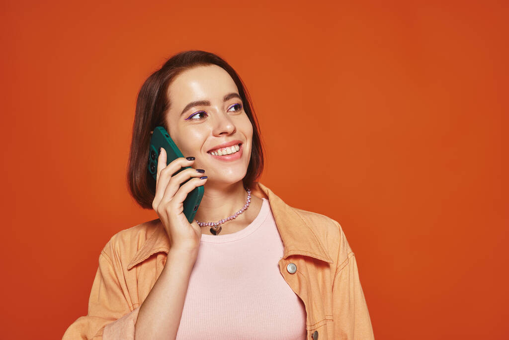 オレンジ色の背景,電話でスマートフォンで話している活気に満ちた服装の幸せな若い女性 - 写真・画像